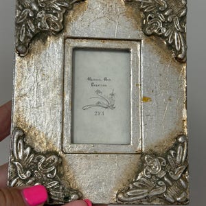 May include: A rectangular picture frame with a silver and gold distressed finish. The frame features ornate corner detailing and a central insert with the text Heather Ann Creations and 2x3 printed in black ink on a light gray background.