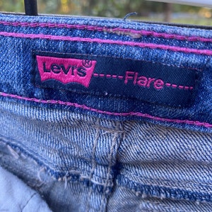 Puede incluir: Un par de pantalones vaqueros azules con detalles de costura rosa. Los vaqueros tienen una etiqueta de Levi's con el texto "--Flare--".