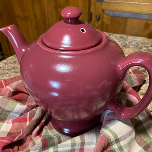 Può includere: Una teiera in ceramica di colore bordeaux scuro con un coperchio e un corpo arrotondato. La teiera è appoggiata su un tessuto a quadri.