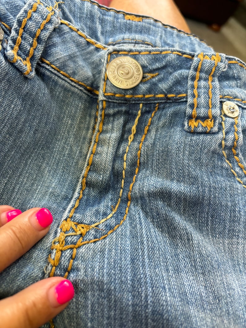 May include: Close-up of light blue denim jeans with gold stitching. The jeans feature a silver button with the text "TRUE RELIGION BRAND JEANS". The stitching is visible along the seams and pockets.