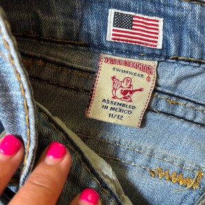 May include: Close-up of blue denim shorts with a small American flag patch. A True Religion Swimwear tag is visible, with text indicating the size is 11/12 and assembled in Mexico. The shorts have visible stitching.