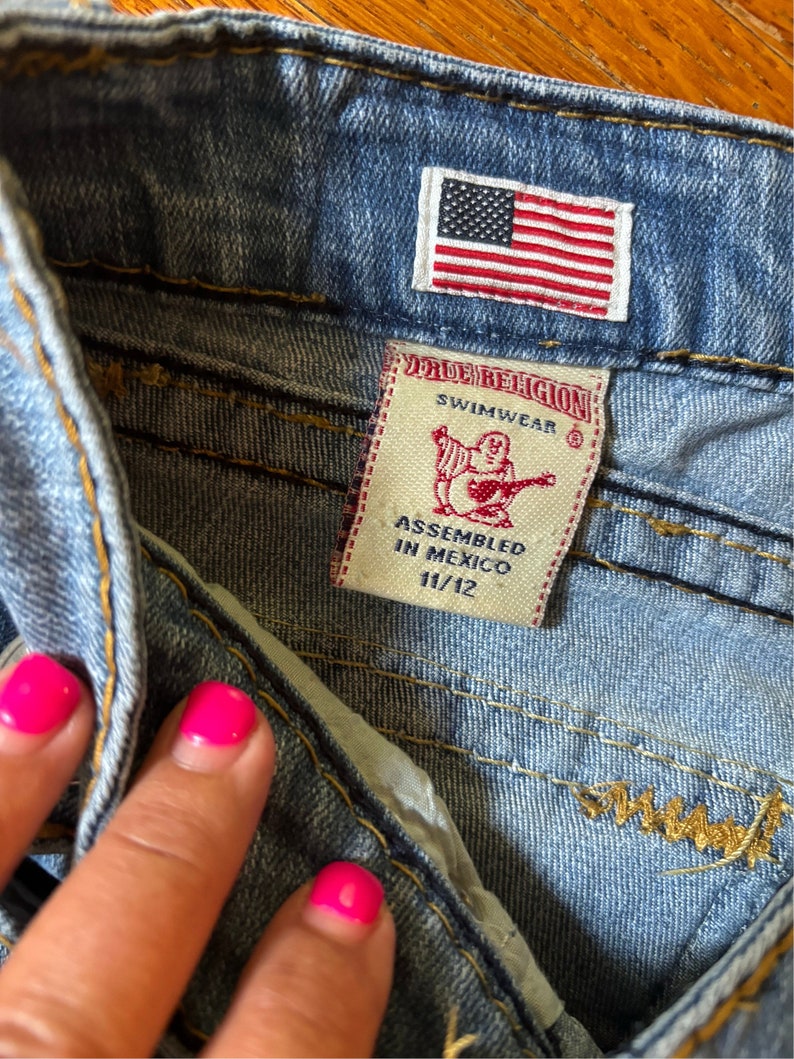 May include: Close-up of blue denim shorts with a small American flag patch. A True Religion Swimwear tag is visible, with text indicating the size is 11/12 and assembled in Mexico. The shorts have visible stitching.