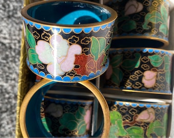 May include: Set of six black and gold napkin rings with blue and green enamel floral designs. The rings are stacked and feature a white flower with pink accents.