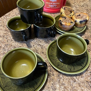 Puede incluir: Cuatro tazas de té negras y verdes con platillos. Los platillos tienen un diseño floral negro y verde. Las tazas de té están llenas de café. Una lata de café roja con el texto "Folgers House Blend" está en el fondo. Hay tres magdalenas de arándanos en un platillo verde.