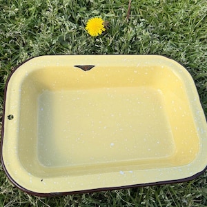 Puede incluir: Una fuente de horno rectangular de esmalte amarillo con un patrón moteado y un borde marrón oscuro. La fuente tiene esquinas redondeadas y un pequeño agujero en un lado. Una flor amarilla está en el fondo.
