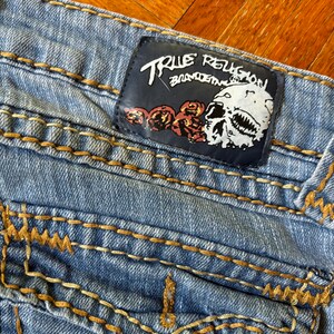 May include: Light blue denim jeans with gold stitching and a leather patch. The patch features the text "TRUE RELIGION" above a skull graphic with roses. The jeans have a classic five-pocket design.