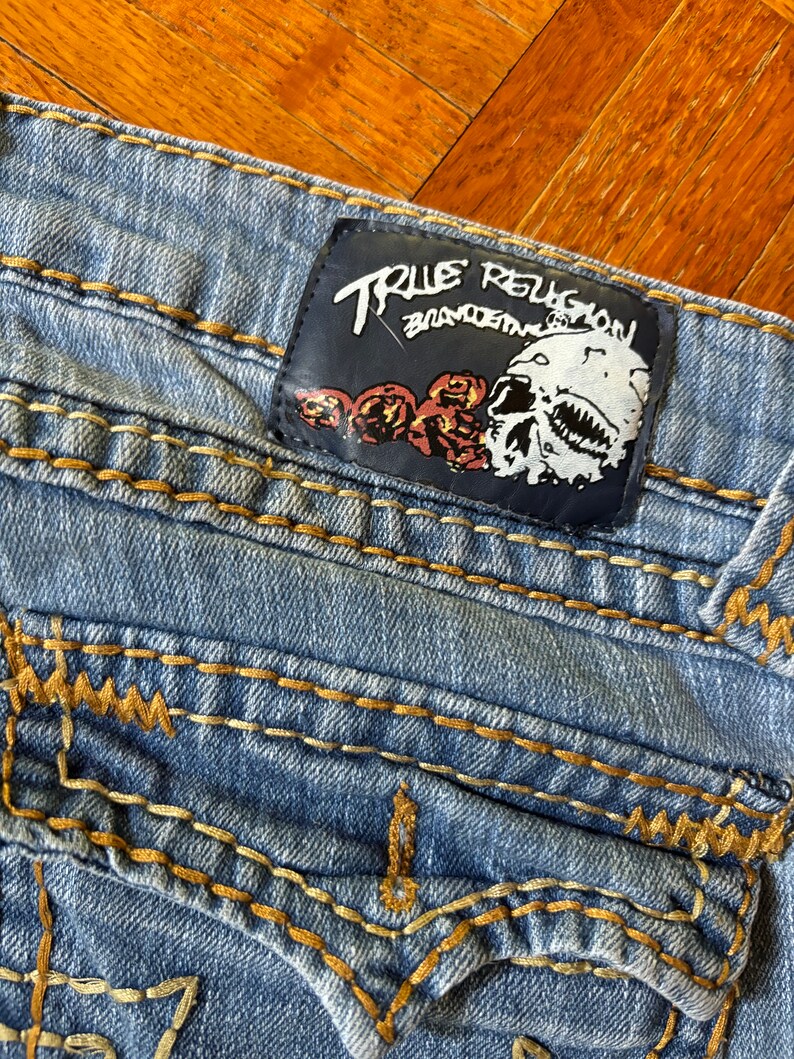 May include: Light blue denim jeans with gold stitching and a leather patch. The patch features the text "TRUE RELIGION" above a skull graphic with roses. The jeans have a classic five-pocket design.