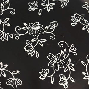 May include: Close-up of a black surface with a repeating white floral pattern. The design features stylized flowers, leaves, and vines, creating a delicate and elegant aesthetic. The pattern is evenly distributed across the surface.