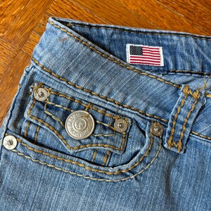 May include: Close-up of light blue denim jeans with a small American flag patch. The jeans feature a pocket with a silver button and decorative stitching in a contrasting gold color. The jeans are made of a durable denim material.