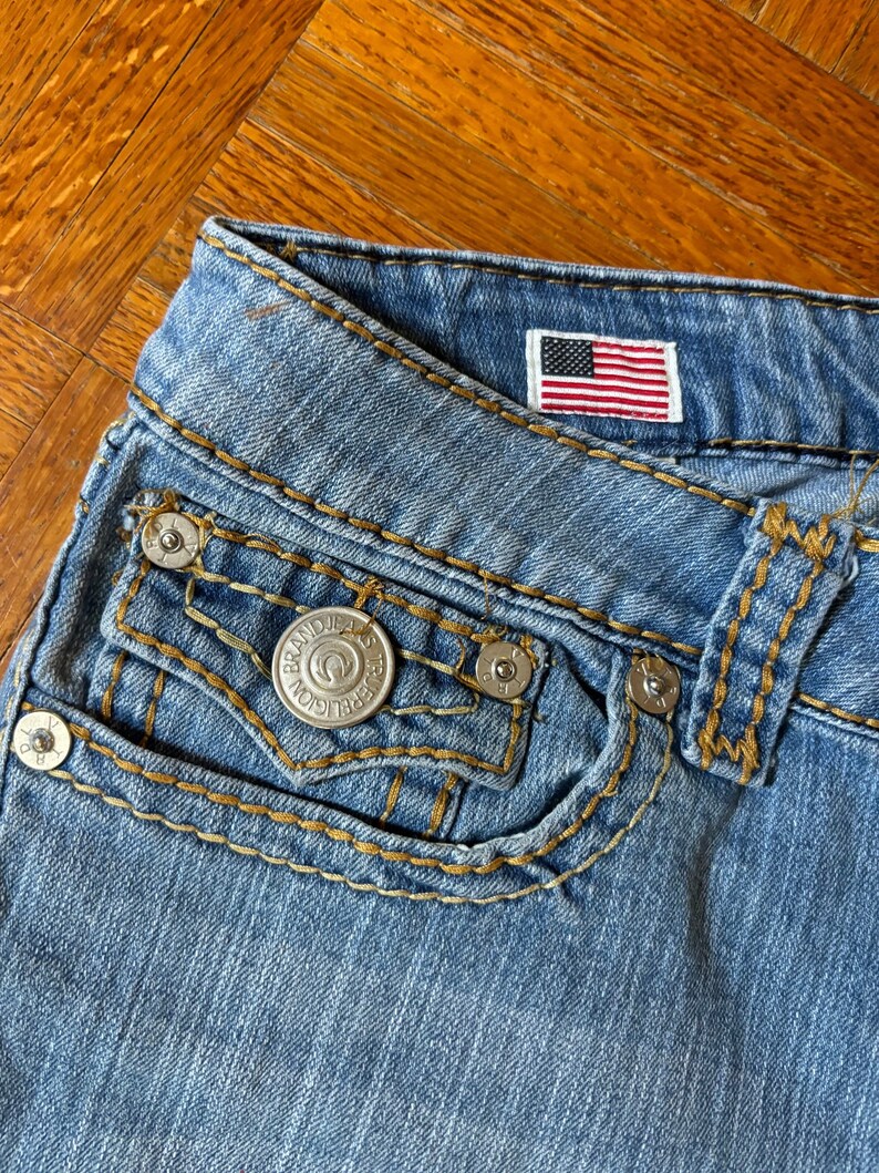 May include: Close-up of light blue denim jeans with a small American flag patch. The jeans feature a pocket with a silver button and decorative stitching in a contrasting gold color. The jeans are made of a durable denim material.
