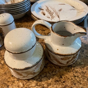 May include: A set of white ceramic dishes with a brown wheat design. The set includes a creamer, a sugar bowl, and several plates.