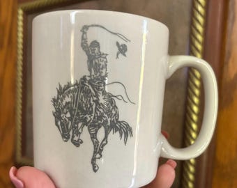 May include: A white ceramic mug featuring a dark gray line art illustration of a cowboy riding a bucking horse. The cowboy is depicted with one arm raised, holding a lasso, while a hat flies through the air nearby. The mug has a smooth finish.