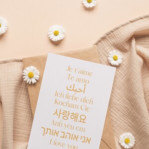 May include: A white card with the words "Je t'aime", "Te amo", "أحبك", "Ich liebe dich", "Kocham Cię", "사랑해요", "Anh y&ecirc;u em", "אני אוהב אותך", and "I love you" written in gold on it. The card is sitting on a brown envelope and a beige fabric.