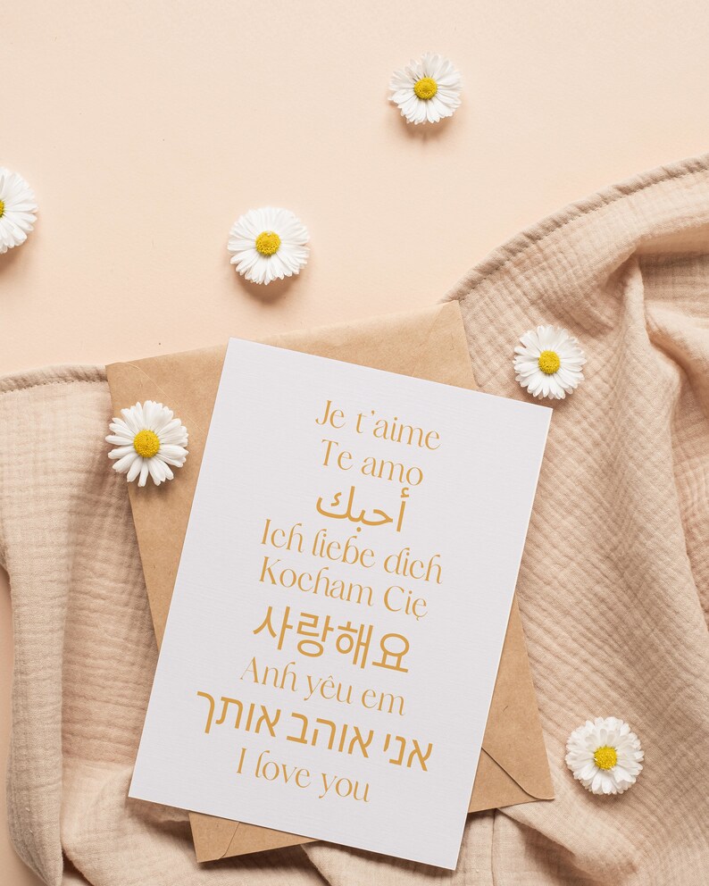 May include: A white card with the words "Je t'aime", "Te amo", "أحبك", "Ich liebe dich", "Kocham Cię", "사랑해요", "Anh y&ecirc;u em", "אני אוהב אותך", and "I love you" written in gold on it. The card is sitting on a brown envelope and a beige fabric.