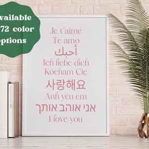 May include: A white framed print with the text "I love you" in pink lettering, written in multiple languages, including English, French, Spanish, German, Korean, Arabic, and Hebrew. The print is displayed on a pink brick wall with a potted green plant in the background.