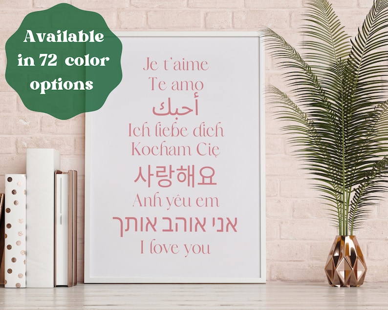 May include: A white framed print with the text "I love you" in pink lettering, written in multiple languages, including English, French, Spanish, German, Korean, Arabic, and Hebrew. The print is displayed on a pink brick wall with a potted green plant in the background.