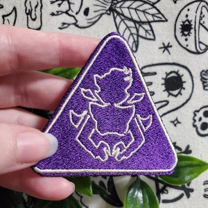 May include: Purple triangular patch with a white embroidered design of a creature with a large head and long arms. The patch has a white border.