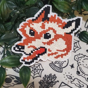 May include: An embroidered patch featuring a pixel art style fox head. The fox is rendered in shades of orange, cream, and black, with a prominent red tongue sticking out. The design has a white border and is shaped to follow the fox's silhouette.