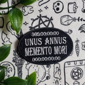 Pode incluir: Etiqueta bordada em preto e branco com o texto "365:00:00:00 UNUS ANNUS MEMENTO MORI 000:00:00:00" sobre um fundo branco com rabiscos em preto e branco.