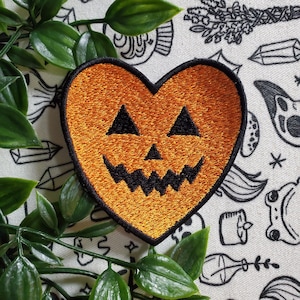 Może przedstawiać: Pomarańczowa, haftowana łatka w kształcie serca z czarną twarzą dyni. Łatka znajduje się na białym tle z czarno-białymi ilustracjami różnych przedmiotów o tematyce Halloween.