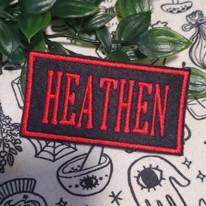 May include: A rectangular black patch with the word "HEATHEN" embroidered in red, framed by a red border. The patch is set against a patterned background with green leaves.