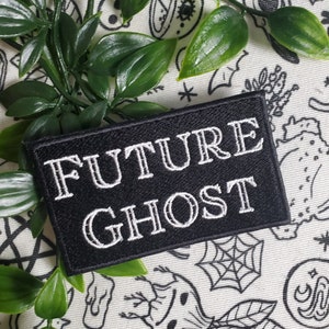 Op de afbeelding: Zwarte geborduurde patch met witte tekst die "FUTURE GHOST" luidt. De patch bevindt zich op een witte achtergrond met zwart-witte illustraties.