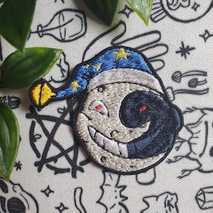 Può includere: Una toppa ricamata che rappresenta una luna sorridente che indossa un cappello da mago blu e giallo. La luna ha un occhio nero e un occhio rosso.