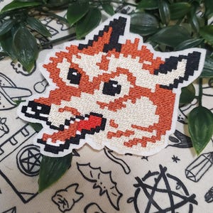 Pu&ograve; includere: Una toppa ricamata raffigurante una testa di volpe in stile pixel art. La volpe &egrave; resa in tonalit&agrave; arancione, crema e nero, con una lingua rosso brillante visibile nella bocca aperta. Il design presenta un bordo bianco e un aspetto squadrato.