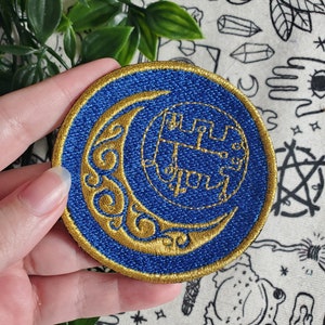 Pode incluir: Um remendo bordado azul e dourado com uma lua crescente com um design estilizado dentro. O remendo é circular e tem uma borda dourada.
