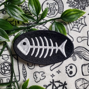 Può includere: Toppa da stirare ricamata in bianco e nero di uno scheletro di pesce su sfondo bianco con scarabocchi in bianco e nero.