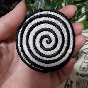 May include: A round, embroidered patch with a black border and a white and black spiral design. The patch is circular and features a detailed, stitched pattern, suitable for application on clothing or accessories.