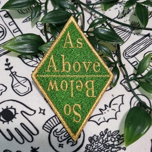 May include: A diamond-shaped embroidered patch with a green background and gold border. The text "As Above" is stitched in gold thread at the top, while the text "So Below" is stitched in gold thread at the bottom, with the word "Below" inverted.