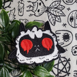 May include: Black and white embroidered patch of a cartoon sheep with red eyes. The sheep has a sad expression and is surrounded by a white fluffy border.