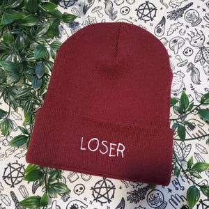May include: A maroon beanie with the word "LOSER" embroidered in white. The hat is made of a soft knit material and is displayed on a patterned surface with green leaves.