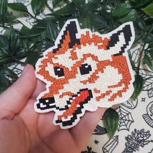 Pu&ograve; includere: Una toppa ricamata raffigurante una testa di volpe in stile pixel art. Il design utilizza filo arancione, bianco e nero per creare i tratti del volto della volpe, incluse orecchie a punta, occhi scuri e un muso con una lingua rossa visibile.