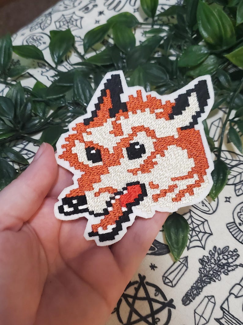 Pu&ograve; includere: Una toppa ricamata raffigurante una testa di volpe in stile pixel art. Il design utilizza filo arancione, bianco e nero per creare i tratti del volto della volpe, incluse orecchie a punta, occhi scuri e un muso con una lingua rossa visibile.
