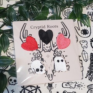 May include: A card with the text "Cryptid Roots" features embroidered patches. The patches include a black heart, two red hearts, a skull, and a ghost. The patches are white, red, and black.