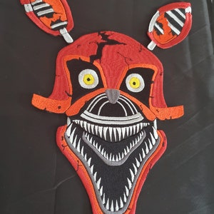 May include: Red and orange embroidered patch of a cartoon character with a large, toothy grin. The character has yellow eyes and a black nose.