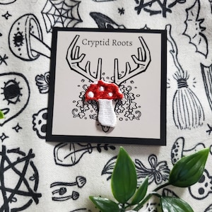 May include: A white card with black text that reads "Cryptid Roots" and a red and white embroidered mushroom with a white stem and a red cap with white dots. The mushroom is on a white background with black floral designs.