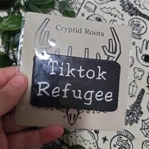Pode incluir: Etiqueta bordada preta com texto branco que diz "Tiktok Refugee" em um fundo branco com uma ilustração em preto e branco de um crânio e chifres. O texto "Cryptid Roots" é impresso acima da etiqueta.