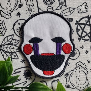 May include: White embroidered patch with a red and black puppet face. The puppet has purple eyelids and a red mouth with white teeth. The patch is on a white background with black and white drawings.