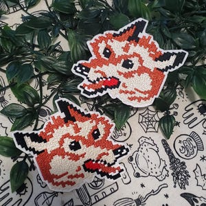 May include: Two embroidered patches featuring a pixel art style fox head design. The fox is rendered in shades of orange, cream, and black thread with a prominent red tongue. Each patch has a white border and a distinct blocky, digital aesthetic.