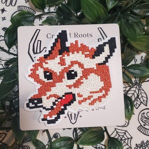 Pu&ograve; includere: Una toppa ricamata raffigurante una testa di lupo in stile pixel art nei toni dell'arancione, bianco e nero. Il lupo ha una lingua rossa visibile ed &egrave; esposto su un cartoncino quadrato con il testo parziale Craft Roots stampato in alto.