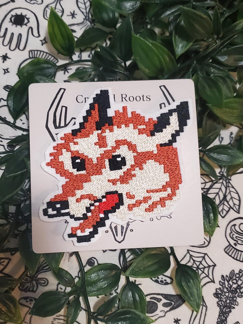 Pu&ograve; includere: Una toppa ricamata raffigurante una testa di lupo in stile pixel art nei toni dell'arancione, bianco e nero. Il lupo ha una lingua rossa visibile ed &egrave; esposto su un cartoncino quadrato con il testo parziale Craft Roots stampato in alto.