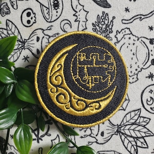 Pode incluir: Um remendo bordado preto e dourado com uma lua crescente e um símbolo dentro. O remendo está sobre um fundo de tecido branco com desenhos em preto e branco de vários símbolos.