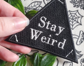 May include: Black triangular patch with white embroidered text that reads "Stay Weird".