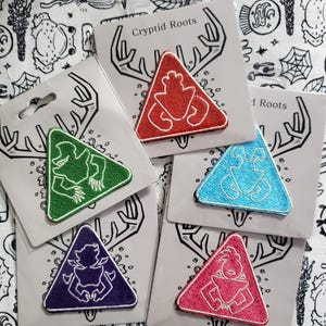 Peut inclure: Cinq patchs thermocollants brodés en forme de triangles. Chaque patch représente une créature différente avec un contour blanc. Les patchs sont sur un fond blanc avec un motif noir et blanc. Le texte "Cryptid Roots" est sur l'un des patchs.