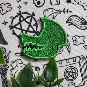 Peut inclure: Patch brodé vert en forme de crâne avec des dents. Le patch est sur un fond blanc avec des dessins au trait noir de divers objets.