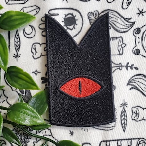 May include: Black felt patch with a red eye in the center. The eye has a black pupil and a white highlight. The patch is shaped like a pointed triangle with rounded edges.