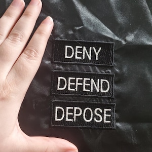 Może przedstawiać: Trzy czarne łatki z tkaniny z białym haftowanym tekstem. Łatki brzmią "DENY", "DEFEND" i "DEPOSE".